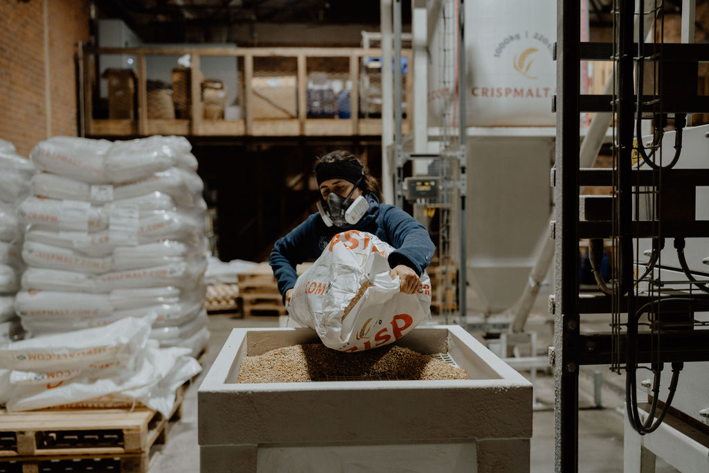 Female distiller, adding peated barley to the milling machine at White Peak Distillery, making Wire Works Whisky